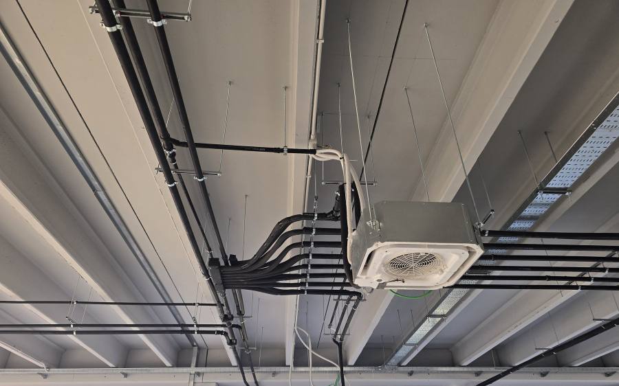 Unità interna a cassetta appesa al solaio a travi di un cantiere al grezzo, con il pannello bianco e la griglia circolare al centro; a sinistra il tubo corrugato bianco della condensa sale in un'ansa e si innesta nella tubazione nera di scarico, mentre intorno pendono i ganci del futuro controsoffitto